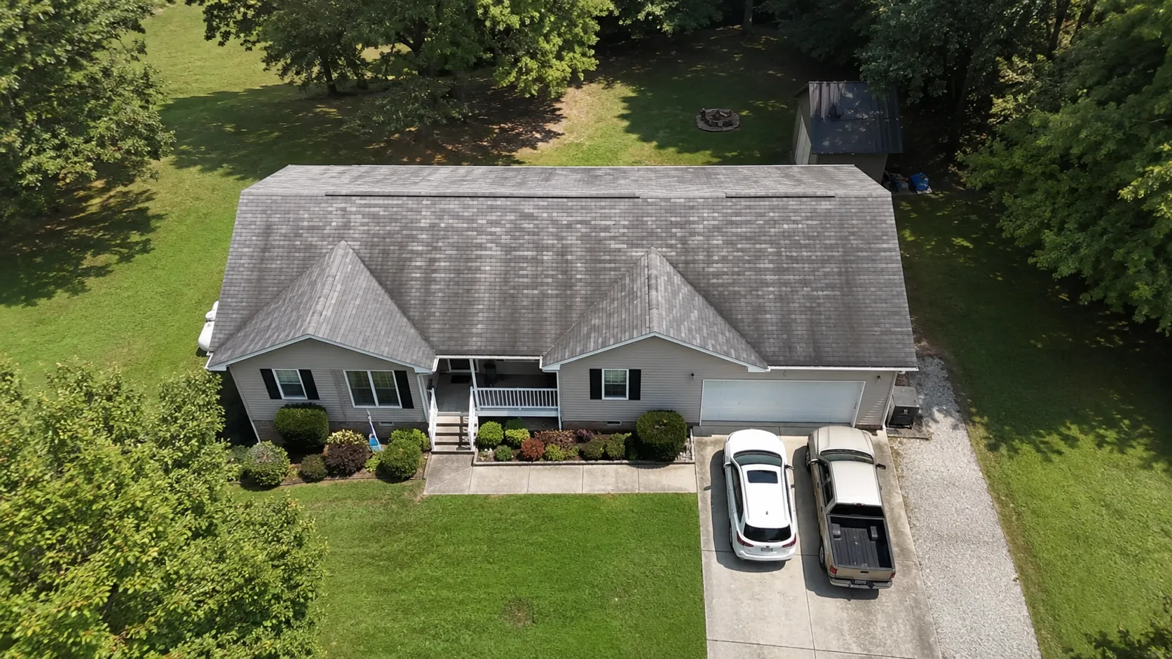 Aerial view of a single-story home with an attached garage and a detached storage shed, roof worn with faded, uneven gray streaking before replacement