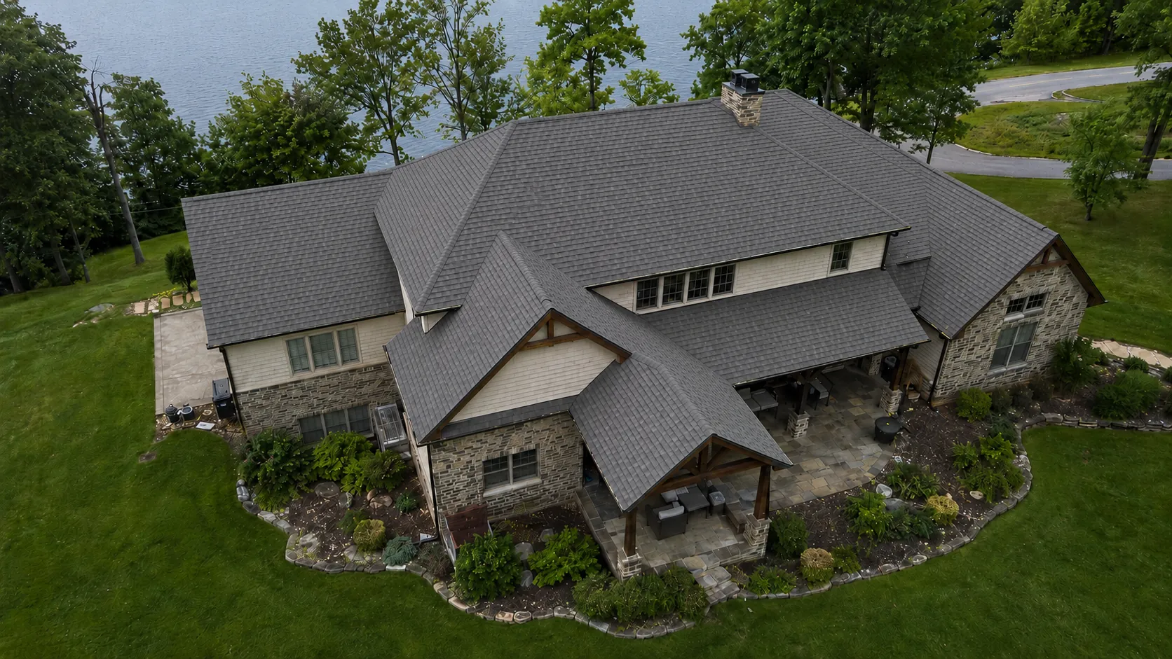 The same lakefront stone and shingle-sided home from above, with its covered patio and chimney, now with a new, uniform gray charcoal architectural shingle roof and straight, even courses