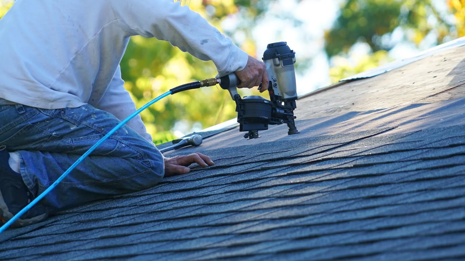 Roofer kneeling on an asphalt shingle roof using a pneumatic coil nail gun, blue air hose trailing behind, with sunlit autumn trees in the background