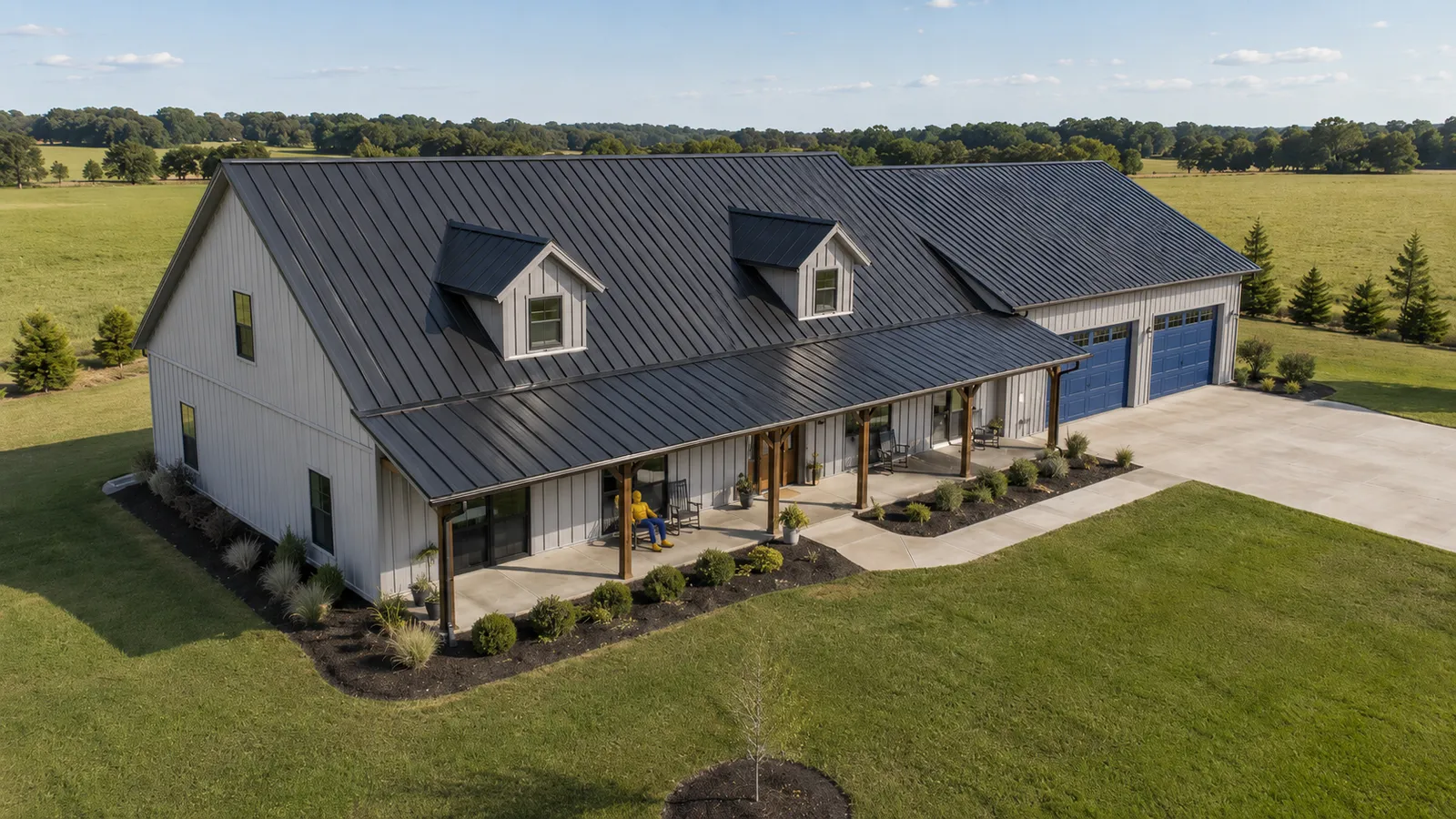 Aerial view of a dark charcoal standing-seam metal roof on a white board-and-batten farmhouse with a covered front porch on timber posts, two dormers, and an attached garage with blue doors, surrounded by green pasture