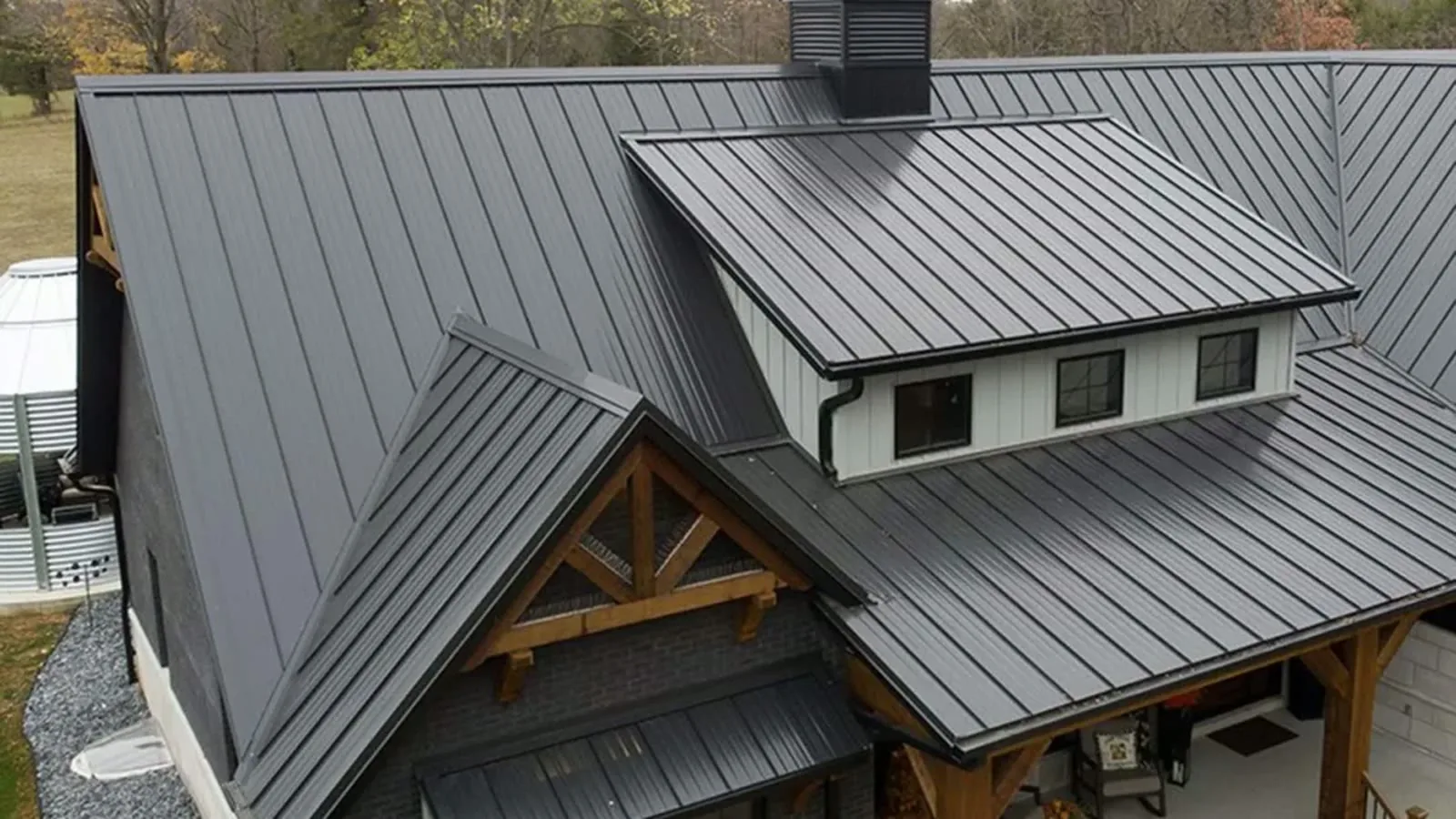Dark standing-seam metal roof with multiple gables, timber-truss porch and cupola on a modern farmhouse