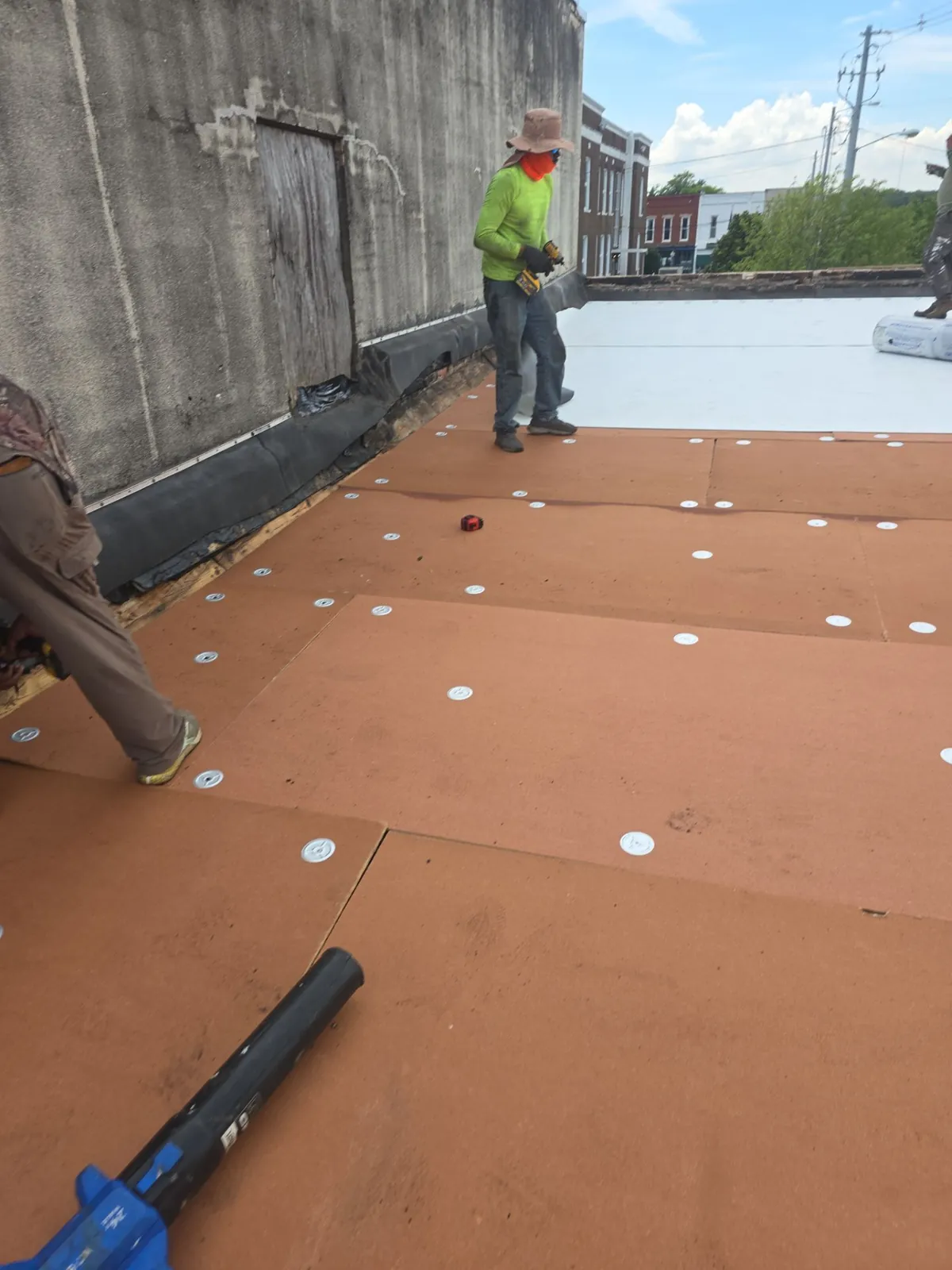 Crew fastening cover board with plates as white single-ply membrane is rolled out behind them