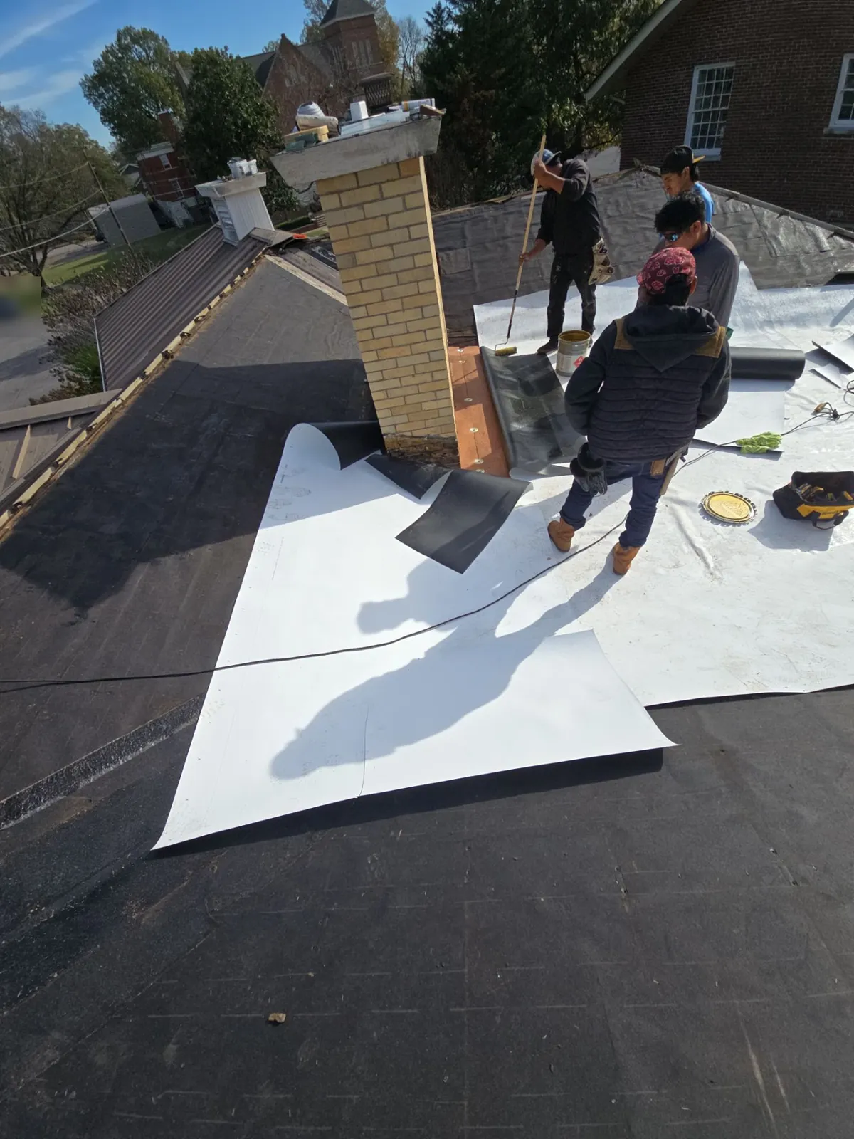 Fit Roofing crew rolling out white single-ply membrane around a brick chimney on a low-slope roof section