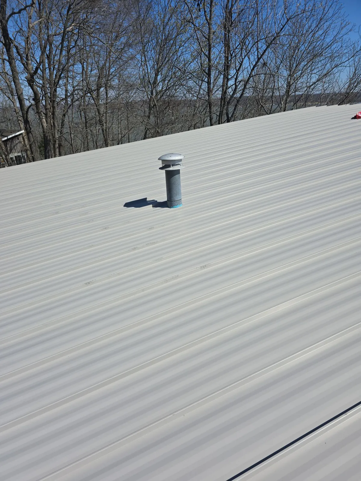 Light gray standing-seam metal panels around a vent pipe, with bare trees behind