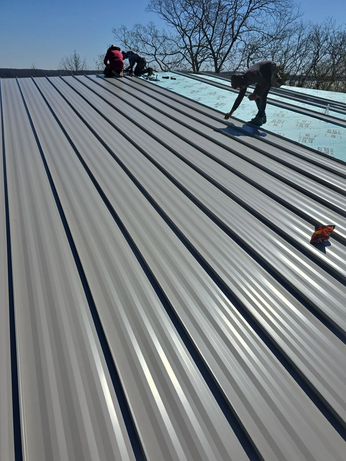 Roofers fastening light gray standing-seam metal panels over synthetic underlayment
