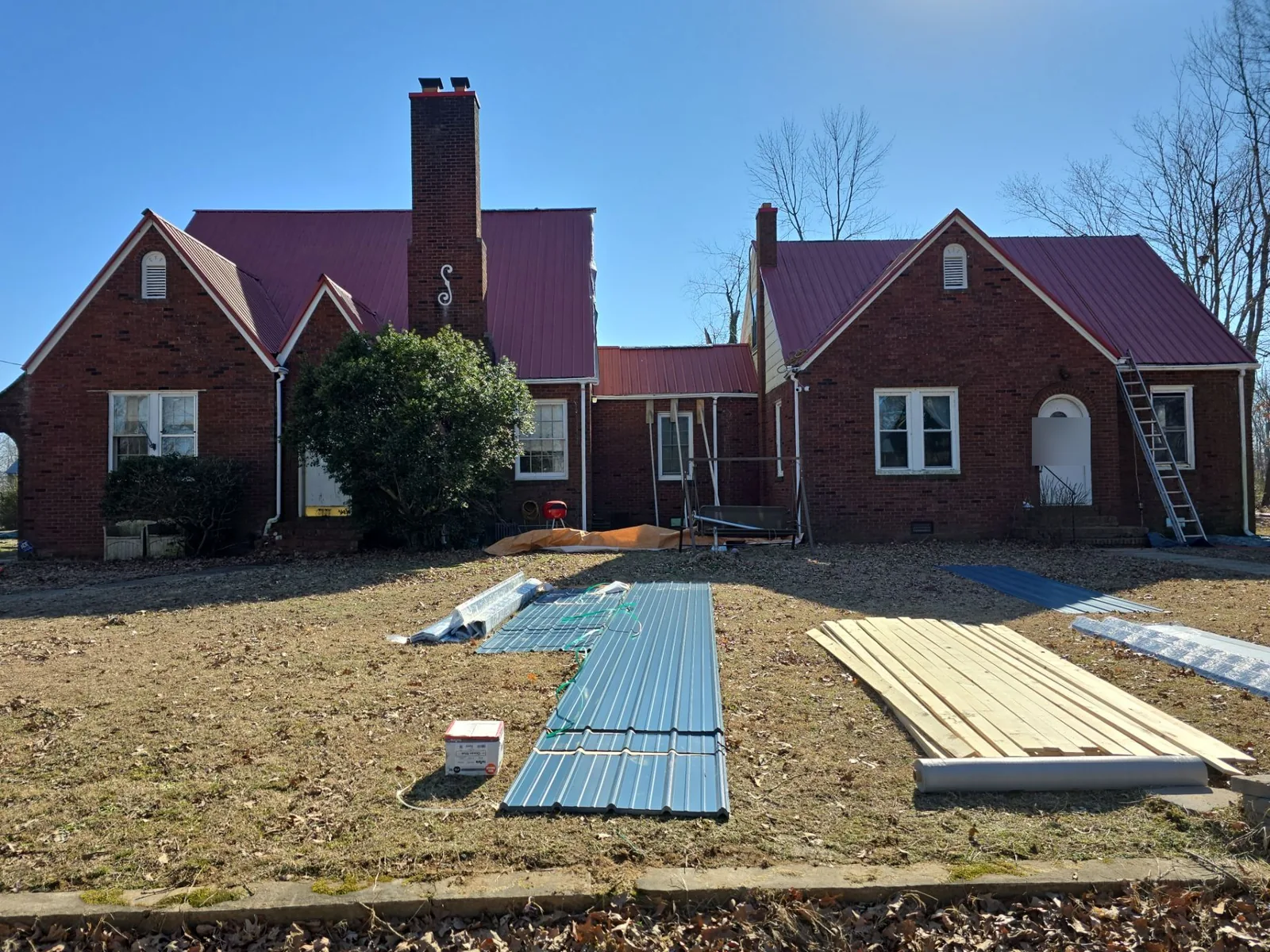 Front of the brick Tudor-style home with its older red metal roof, with new blue metal panels and lumber staged in the yard