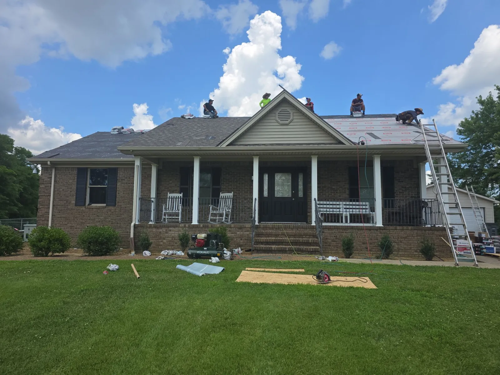 Fit Roofing crew on the roof of the brick ranch home as new underlayment goes down, with ladders and materials in the yard