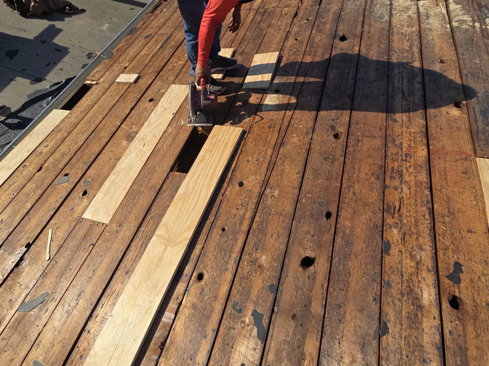 Old plank roof deck exposed at tear-off, with worn boards and holes, as a roofer cuts in new boards