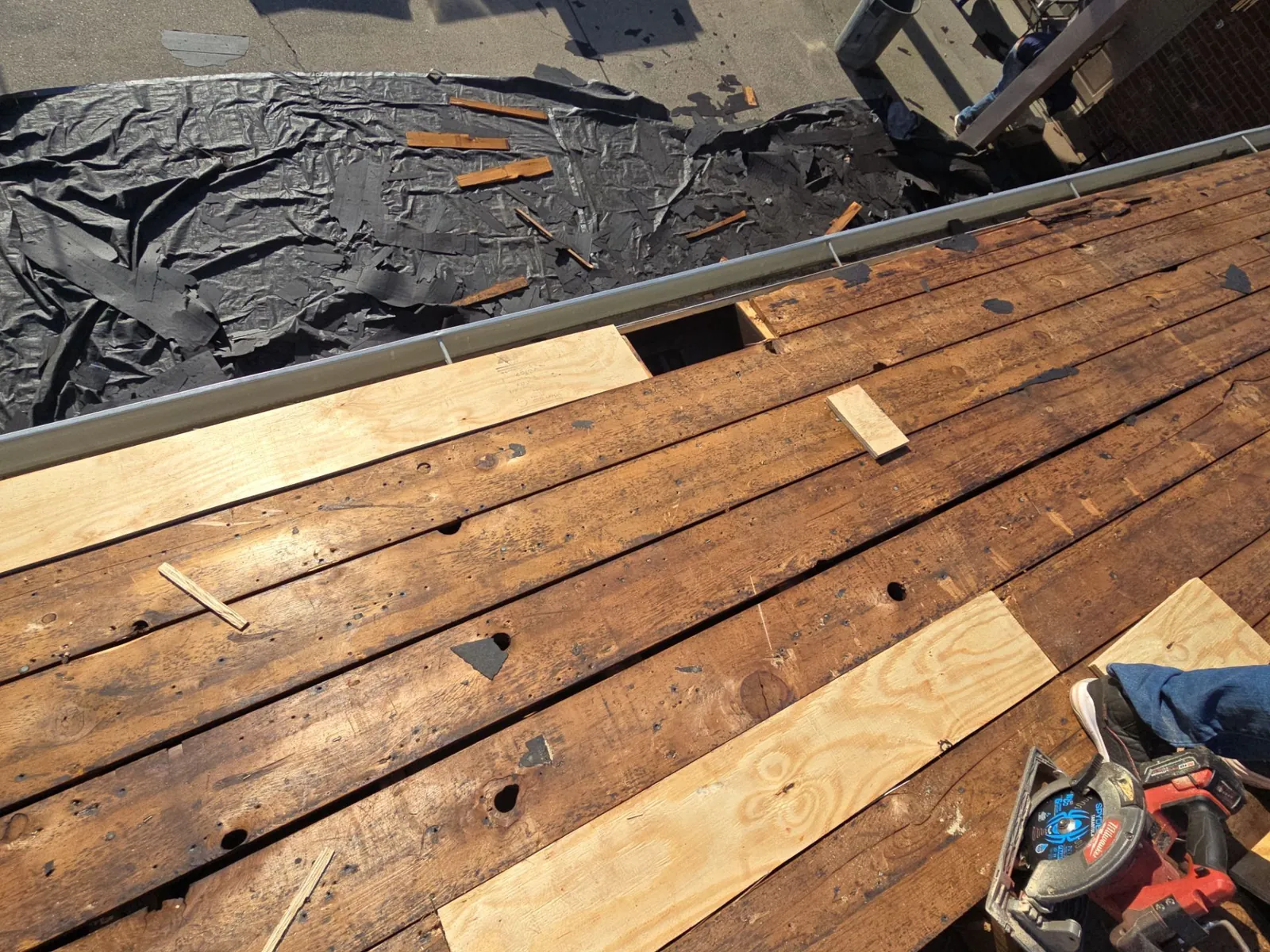 New boards and plywood patched into the old plank deck near the gutter, with tarps laid below to catch debris