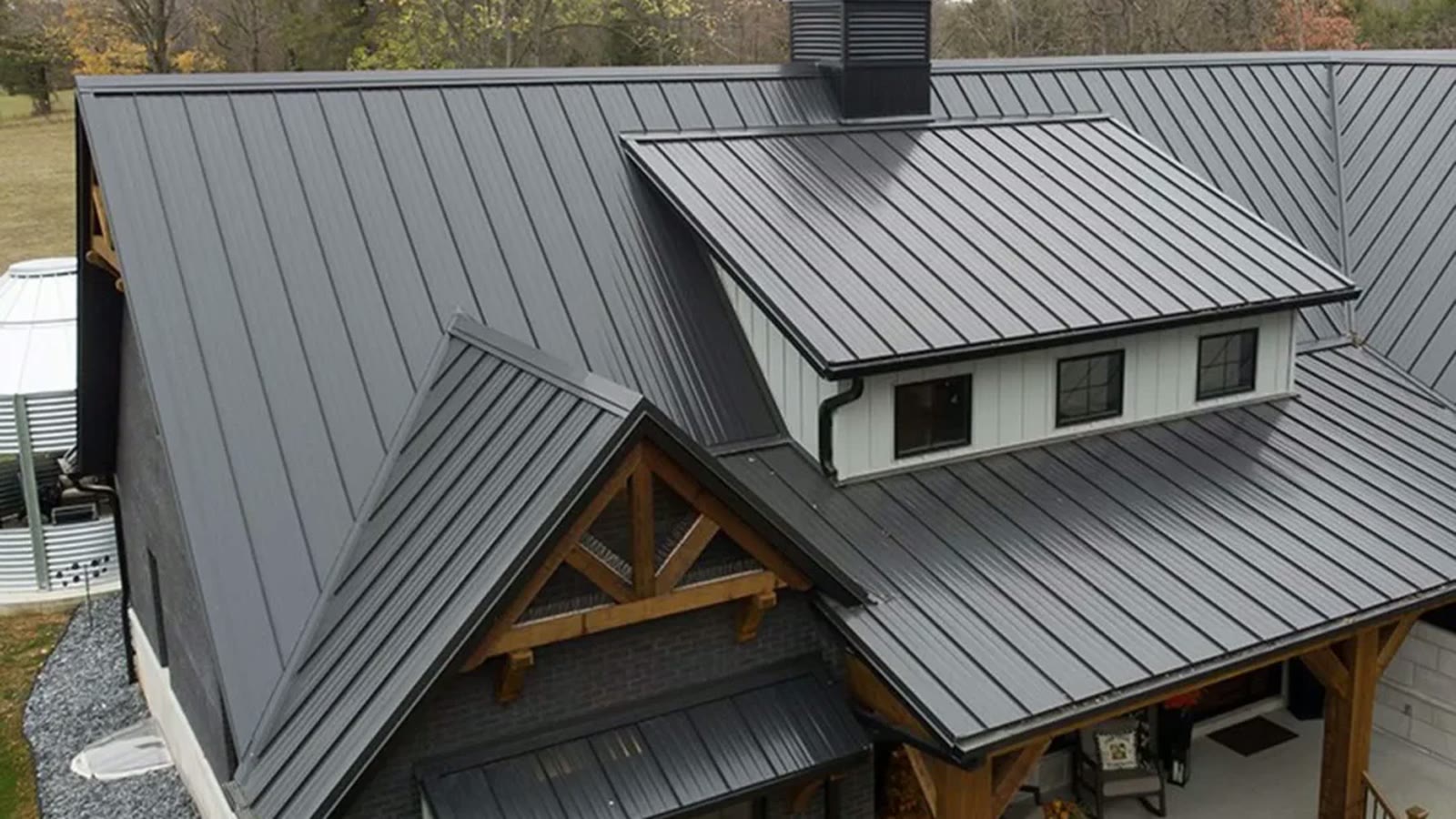 Dark standing-seam metal roof with multiple gables, timber-truss porch and cupola on a modern farmhouse